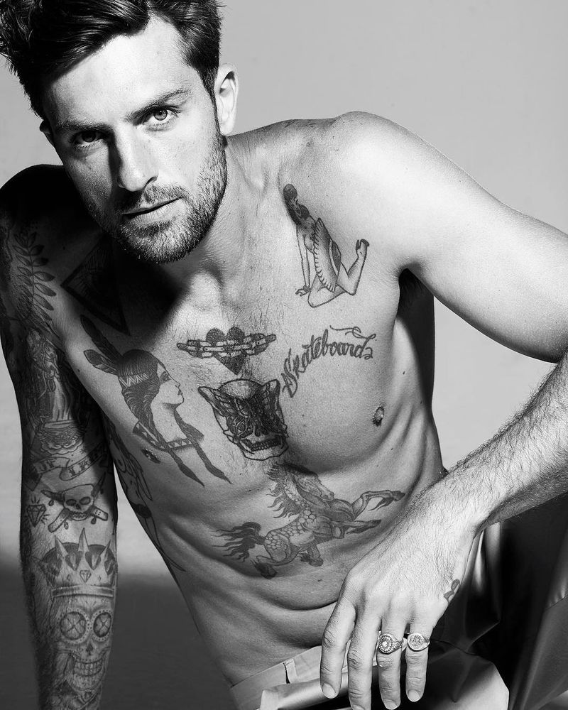 Black and white photo of Brazilian man seated on the floor facing the camera and cropped to upper body and hips. He has short hair and a short beard and a serious expression. His right arm and chest have multiple tattoos of people and objects in a traditional style. His left elbow is rested on a raised knee and rings are visible on his little and ring finger.