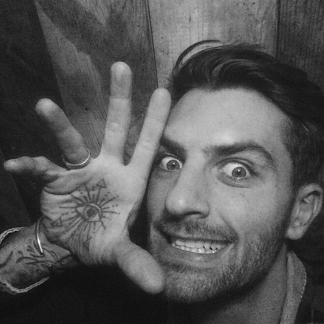 Colour photo of Brazilian man facing towards the camera and cropped to his head. He has short hair and a short beard. His hand is lifted beside his face with an eye tattoo on his palm visible and fingers spread. Is is smiling with a somewhat goofy expression.