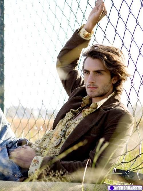 Colour photo of a Brazilian man cropped to upper body; he is half sitting-half laying against a wire fence on the right, with legs out of frame to the left. He is wearing faded blue jeans, a cream floral shirt, and brown jacket. He has chin-length hair and a light beard. His right arm is lifted and holding on to the fence above his head. He is looking to the left with a neutral expression