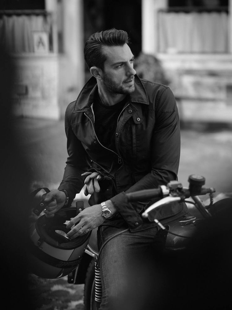 Black and white photograph of Brazilian man sitting astride a motorbike; the photo is cropped to focus on the man from head to knee. He has short hair and a light beard. He is looking to his left but body is twisted to the right with hands holding his helmet. He is wearing jeans, a jacket, and a watch on his left wrist. There are gloves in his right jacket pocket with the fingers poking out.