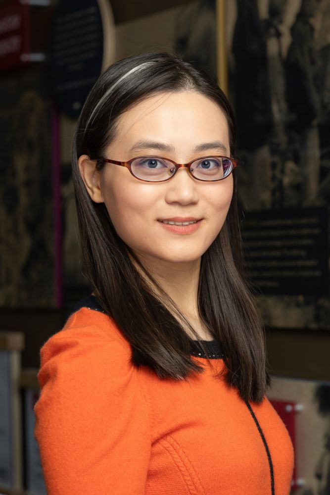 Weidi Qin wearing an orange sweater and glasses standing in a school hallway.