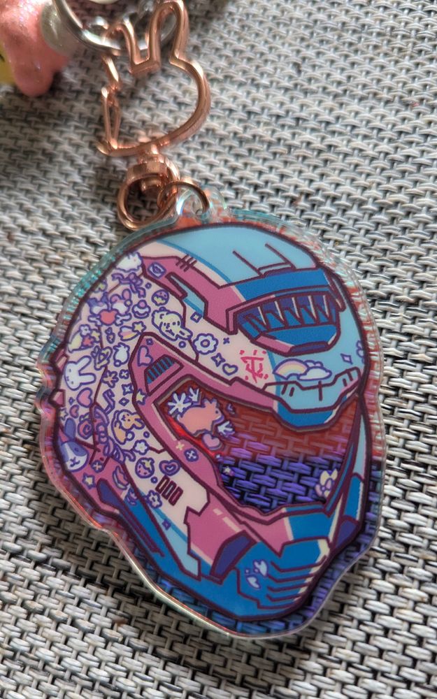 Photo of a keychain, shaped like the doomsayer's helmet. It is colored in the trans flag colors, and covered in stickers, mostly rabbits and bunnies. The keychain also sports a clasp that is shaped like a bunny head. The acrylic is see through, making the visor portion of the helmet clear, with a slight holographic sheen to it that can be seen in the photo. Design by Sanchomps. 