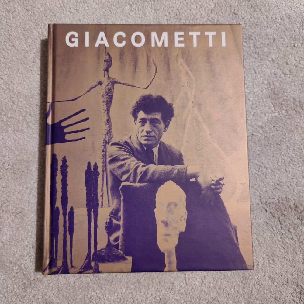 Front cover of book. Giacometti. Full-cover image of Giacometti sitting down, surrounded by his sculptures of tall, thin people. 