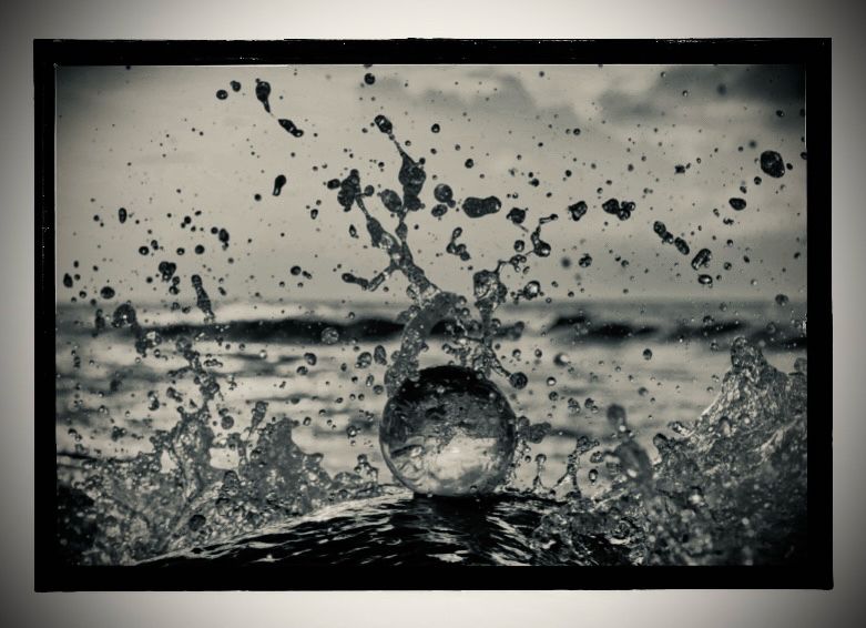 Ein schwarz weißes Foto, auf dem in der Mitte eine (Glas-) Kugel auf einer Welle im Wasser zu sehen ist, die vielleicht ins Wasser fallen gelassen wurde, da um sie herum das Wasser in sämtliche Richtungen spritzt. Im Hintergrund zu sehen Wellen und etwas wolkigen Himmel. 