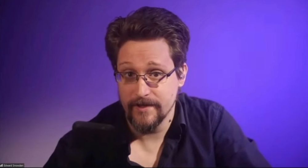 Screenshot from the video clip with a portrait of Julian Assange speaking in front of a neutral violet background.