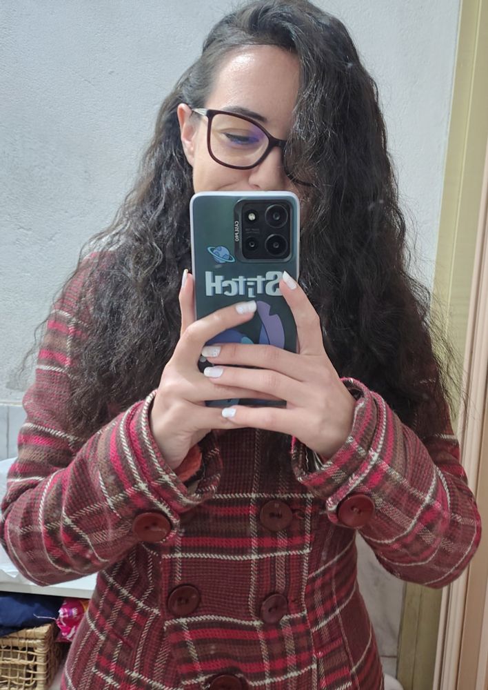 Mirror selfie of woman wearing glasses and a red plaid coat, she has curly brown hair and white nails and her phone has a Stitch case. 