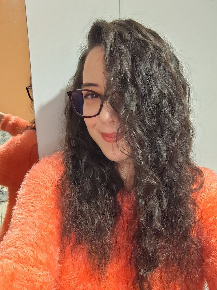 Selfie of a woman with curly brown hair wearing glasses, red lipstick and an orange fuzzy sweater.