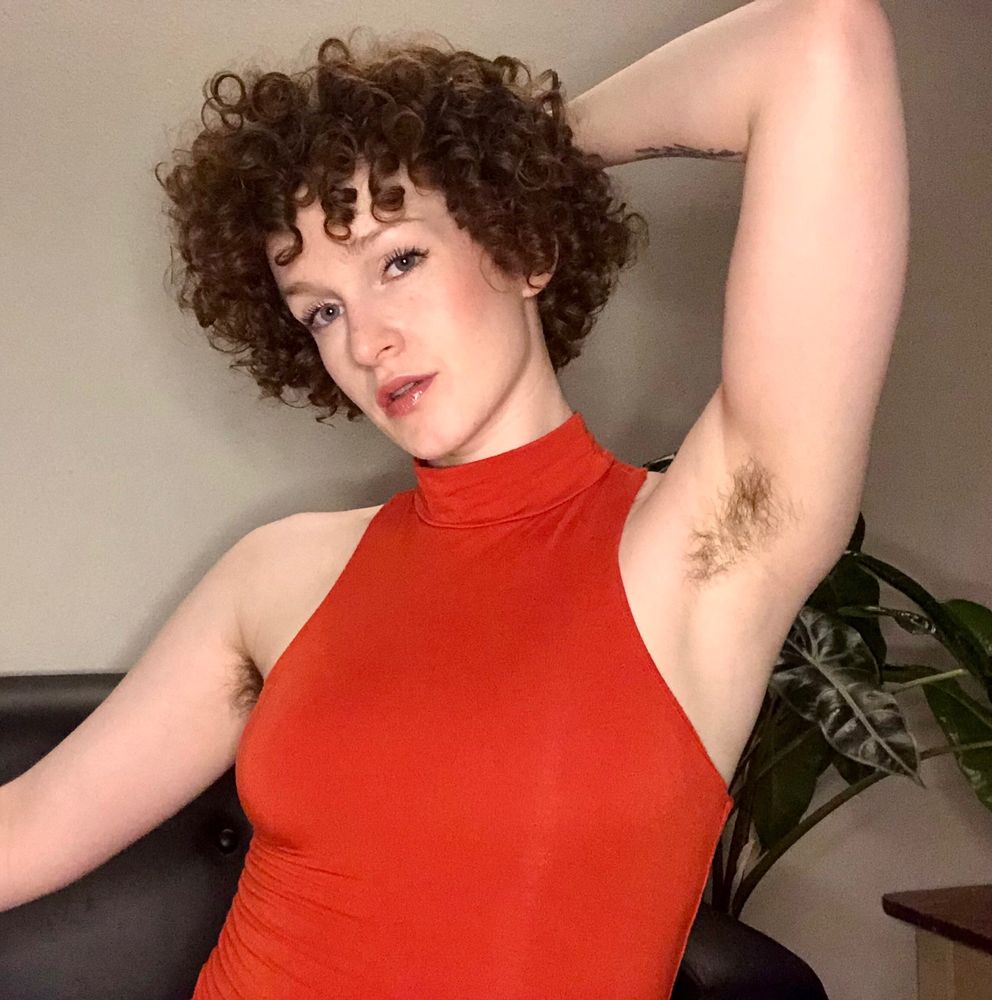 Goddess Diana is in a tight orange turtleneck dress. They are showing their sweaty armpit off to the camera with a gentle smile on their face.