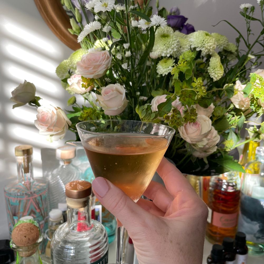 A hand with pale pink painted nails holds a martini glass in front of a vase of flowers including small pale pink roses. 
