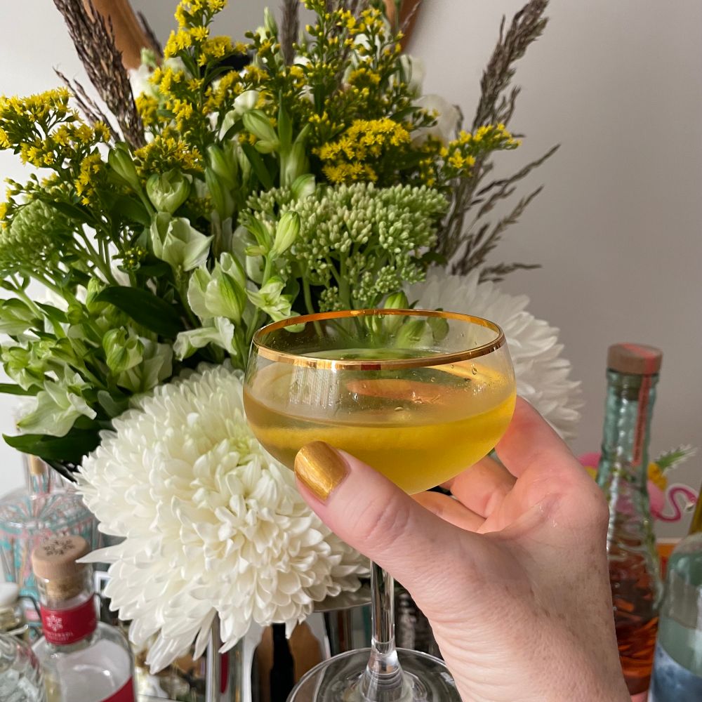 A hand with gold painted nails holds a gold rimmed cocktail glass containing a yellow liquid in front of a vase of yellow and white flowers. 
