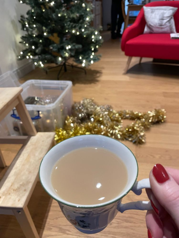 A hand with red painted nails holds a china cup of tea. Behind it on the floor is a pile of gold tinsel. Behind that is a lit up but undecorated Christmas tree. 