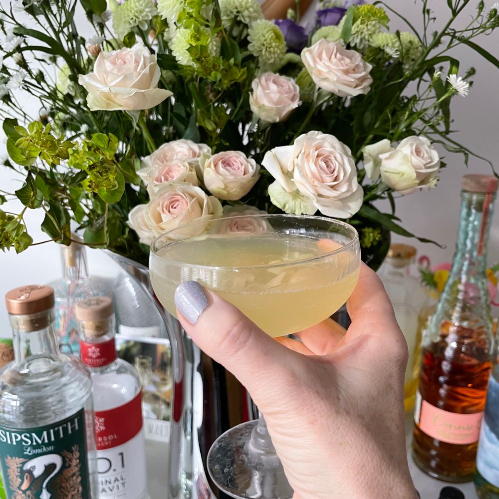 A hand with silvery pink painted nails holds a cocktail glass containing a lemon yellow liquid in front of a vase of flowers including small pale pink roses. 