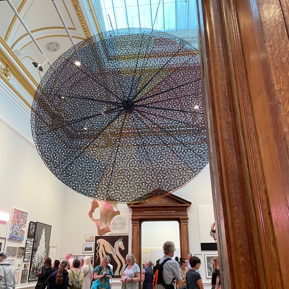A view of a gallery with lots of paintings, and a large pierced disk hanging from the ceiling. 