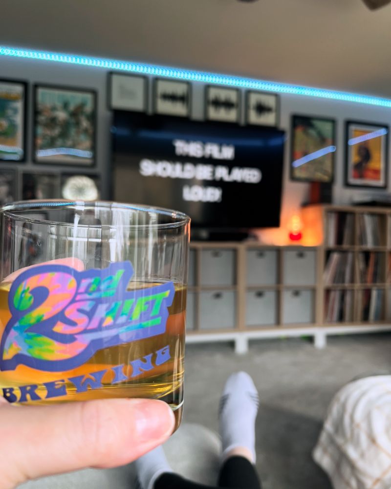 A glass with a beverage and a screen behind the glass that says “this film should be played loud”