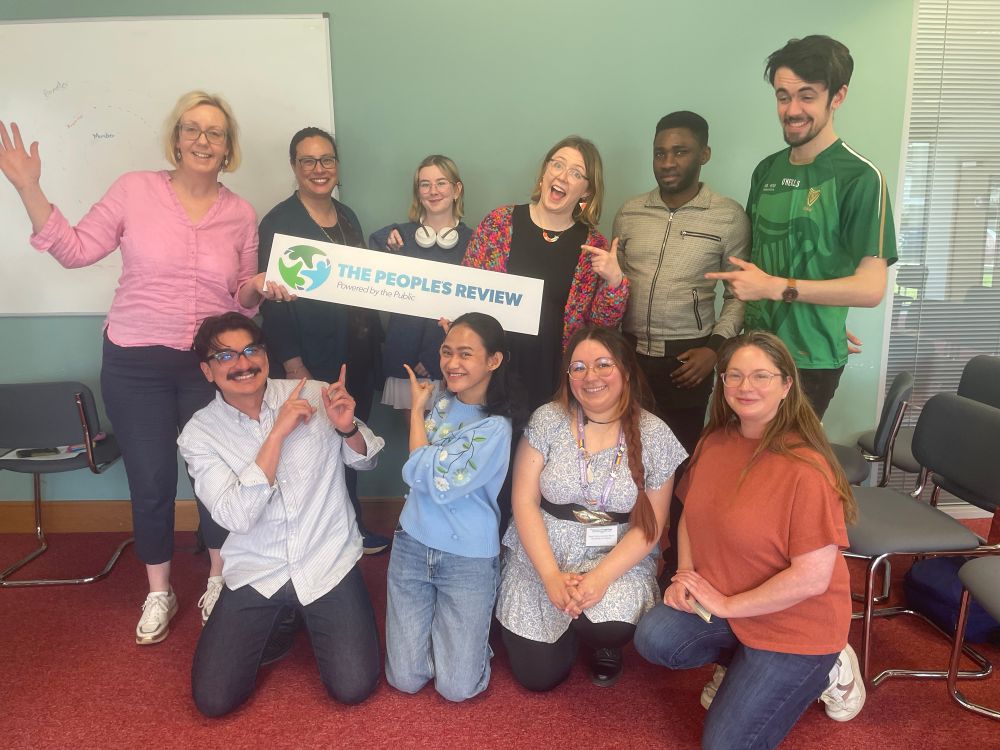 A picture of 10 happy humans pointing at a sign saying The People’s Review - Powered by the Public 