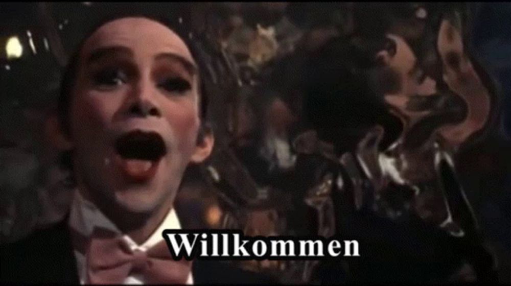 a man in a tuxedo with the word willkommen on the bottom right