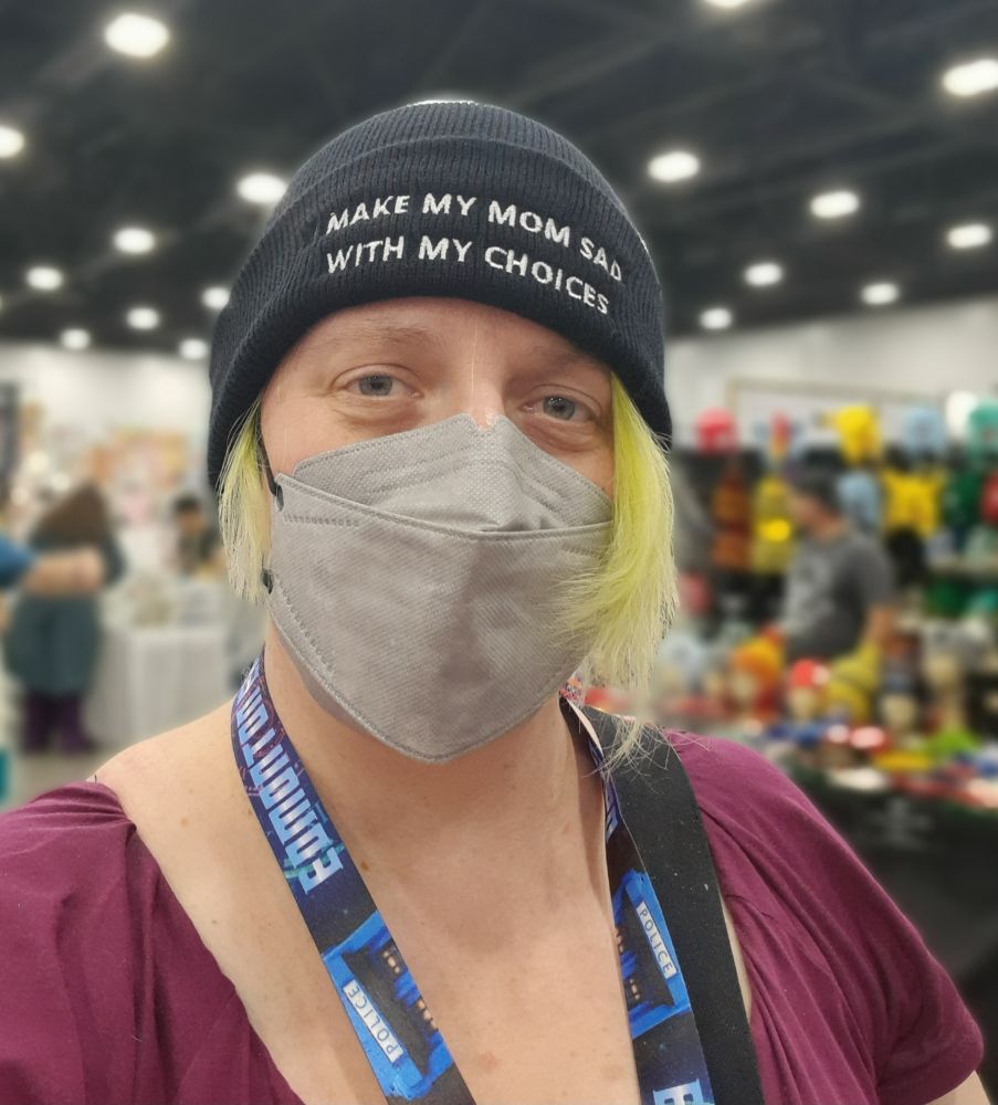A nonbinary person with green hair and a grey face mask wears a beanie that says "I make my mom sad with my choices" 