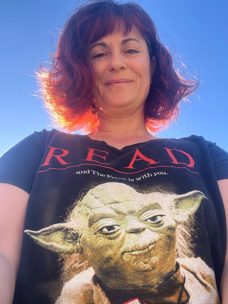 Photo of a white woman with red hair, backlit by the Sun, displaying her T-shirt which says: Read, and the force is with you. The T-shirt features a photo of Yoda from the Star Wars films.