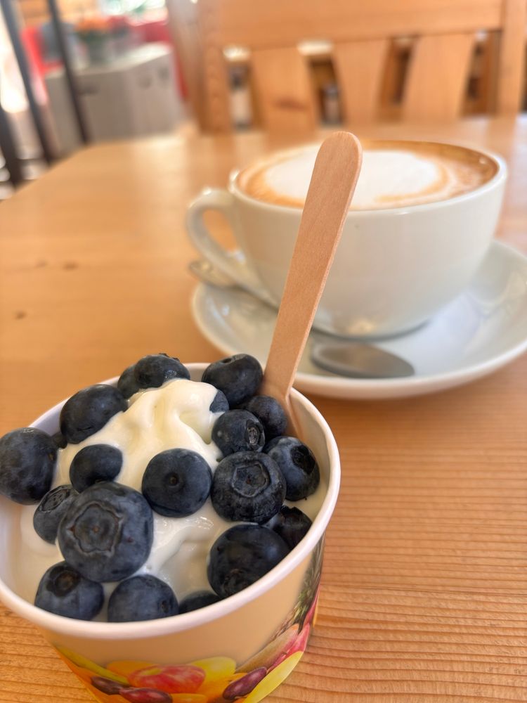 Im Vordergrund auf einem Holztisch ein kleiner Becher frozen Joghurt mit Joghurt, im Hintergrund ein Cappuccino. 