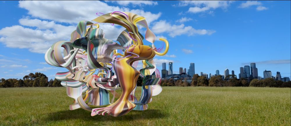 An augmented reality view of a digital sculpture, of spherical form, comprising multicoloured wavy pieces,  and its shadow, in an expanse of open grassland with trees and a city skyline in the distance. A building in the distance has a wavy form on its facade. The sky is blue with some bright clouds.