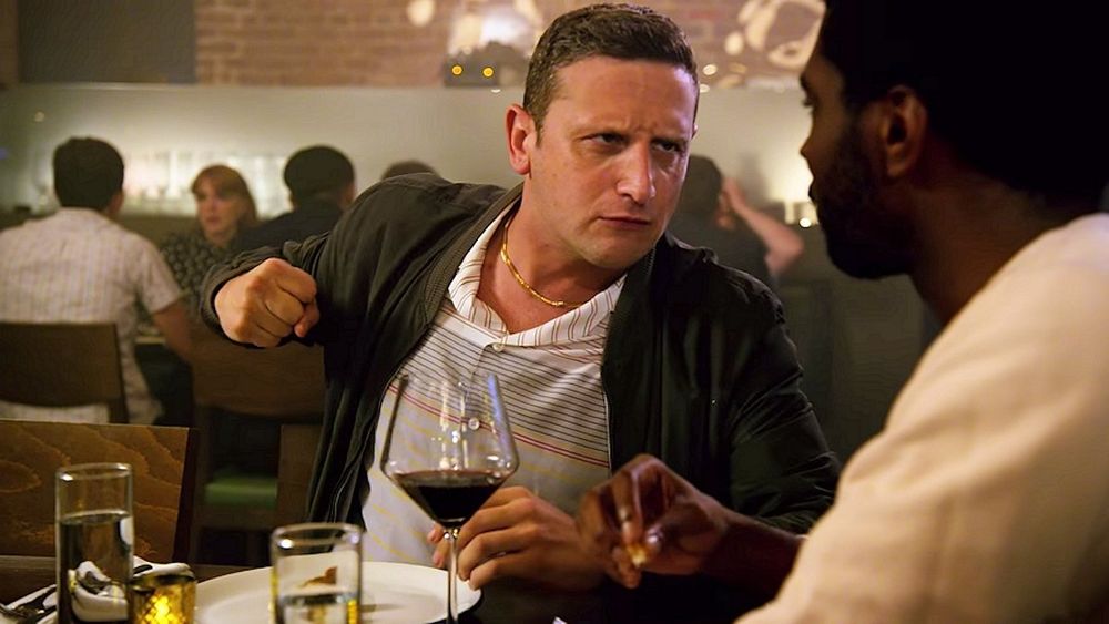 A man, pretending not to be choking on some food, cocking his fist at his friend in a threatening manner for suggesting that he seek medical attention for choking on some food.