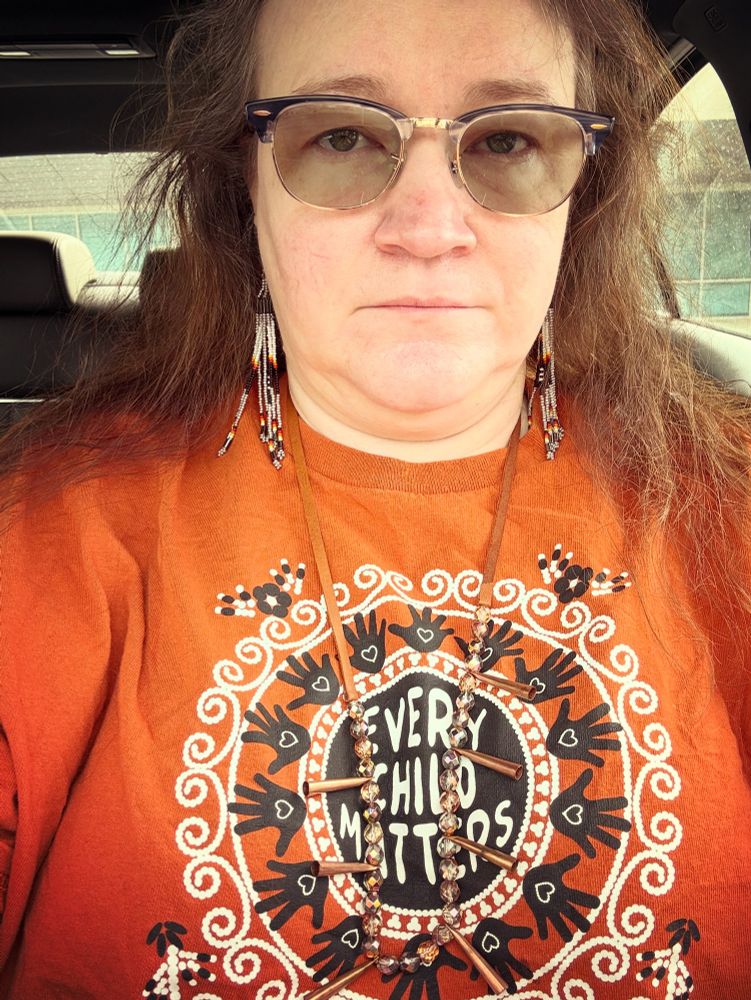 A person with long greying brown hair and glasses wears an orange T shirt with Haudenosaunee geometric and floral designs in black and white and a black central circle labeled EVERY CHILD MATTERS in honor of the commemoration of generations of children lost to Indian boarding school genocide across the USA and Canada.  