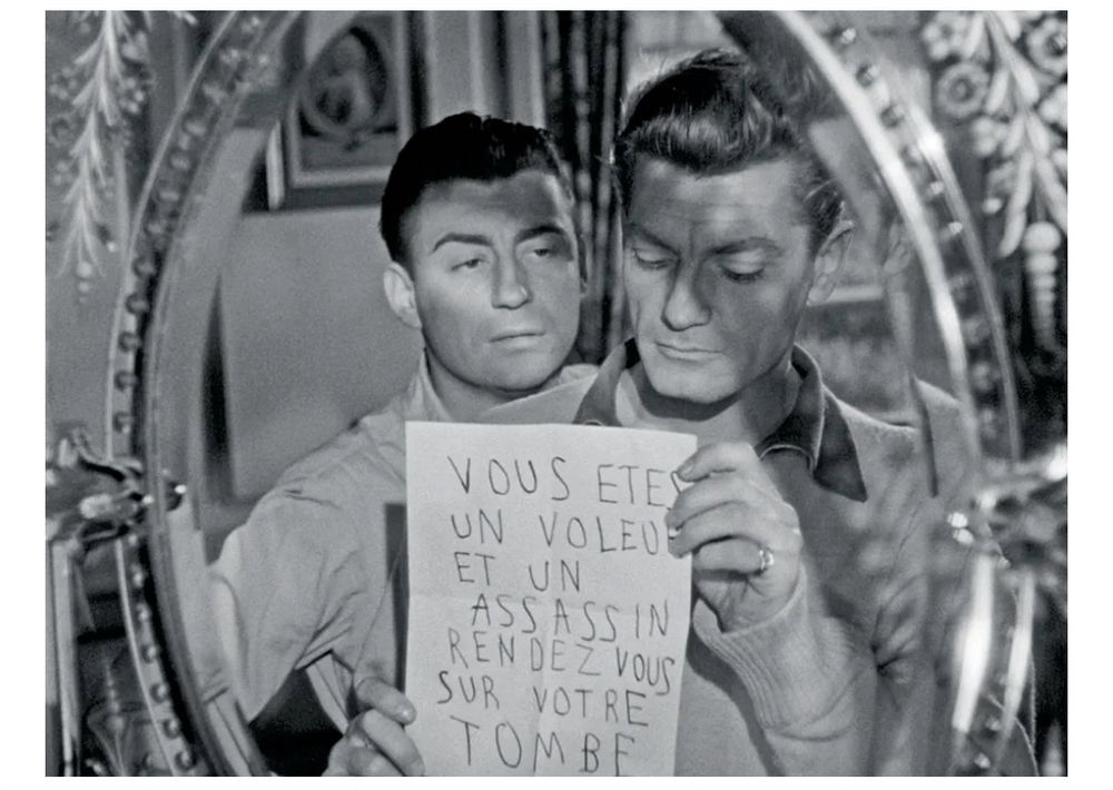Scene from black and white film of a man holding a reversed message in French in the mirror that says “vous êtes un voleur et un assassin rendez-vous sur votre tombe.” 