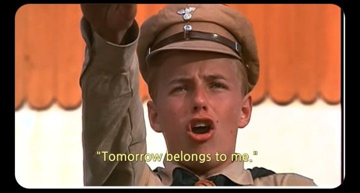 young blonde man with a brown uniform and hat with a nazi insignia. he is raising his arm in a seig heil salute and singing 