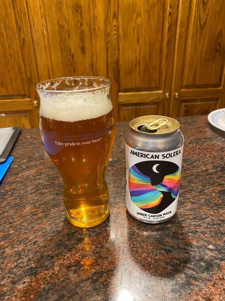 Glass of beer with a can next to it. The can  is white with a black circle graphic containing rainbow hills & a sliver moon