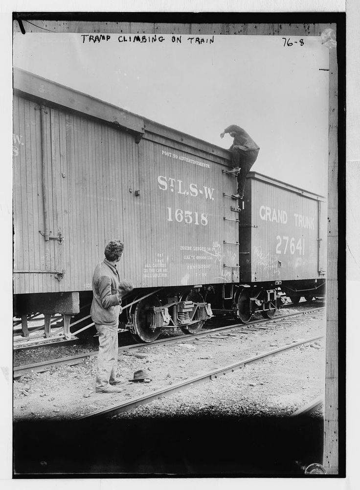 Tramp climbing on railroad car, another man standing between tracks and looking up to watch, hat on ground. Bain News Service, publisher