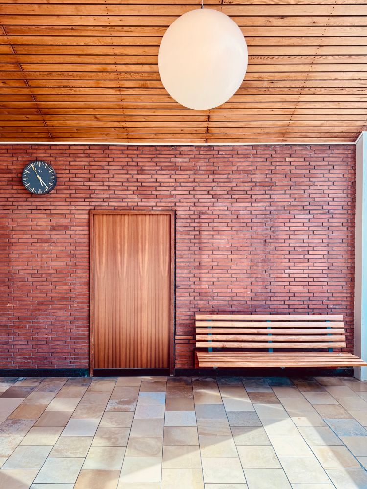 A wooden bench and wooden door stand in contrast with a red brick wall, stone floor and wooden ceiling. To the left of the door hangs a black clock.