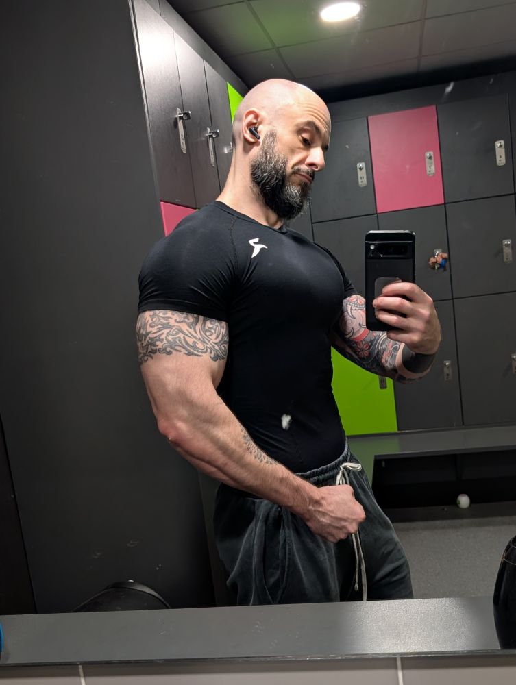 Deviir mirror selfie. Black compression t-shirt. Left hand down by waistband, flexing arm. Chest/pecs pumped