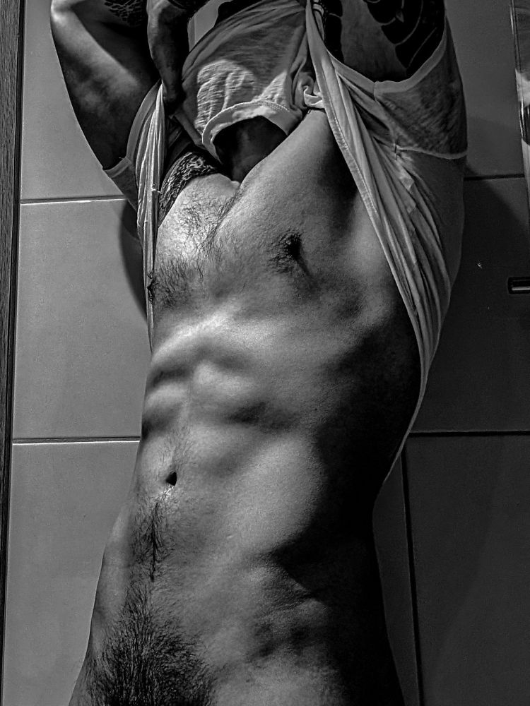 Deviir pulling a wet t-shirt above his head, naked underneath but photo cropped just above crotch. Black and white filter.