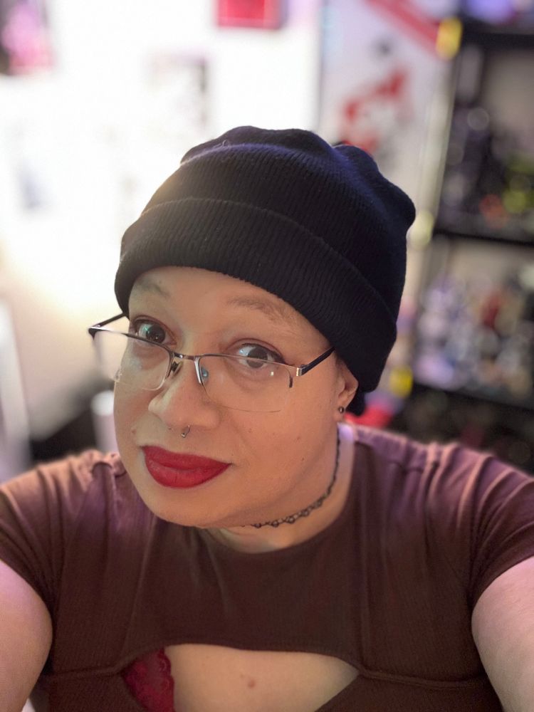 xera selfie

xera wearing a black beanie rectangular glasses, and stretchy choker with a red lip and a brown shirt with a window a hint of a red lacey bra peaks through

A blurred light is behind her 

Her earrings are small black studs