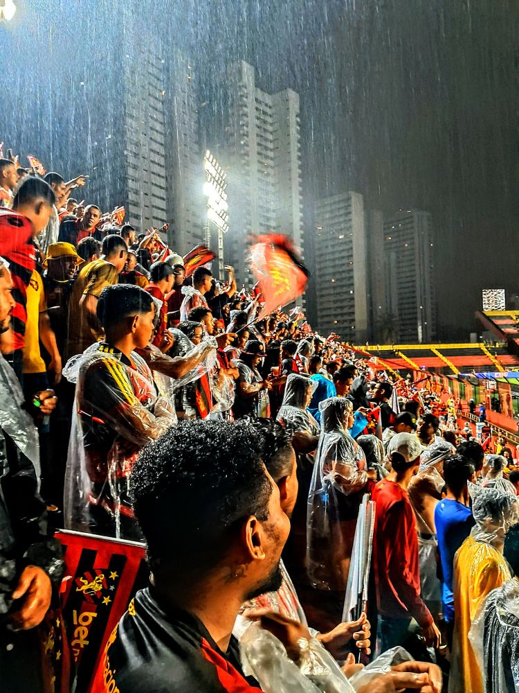 Torcedores do Sport Club do Recife com bandeiras do time, na arquibancada do estádio da Ilha do Retino assistindo ao jogo enquanto a chuva cai.