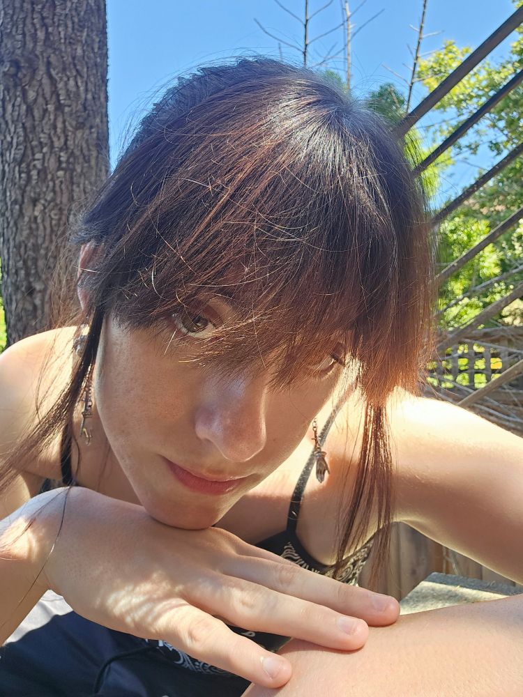 GoblinGirl basks in the sun, bends, and leans her chin on the back of her hand. Her black hair is up in a ponytail with her bangs messily all over the place. in some parts, the dye is fading to brown. she is barefaced and holding a neutral expression while staring thru the camera and at you. her crop top shows off her smooth shoulders and soft skin. 
