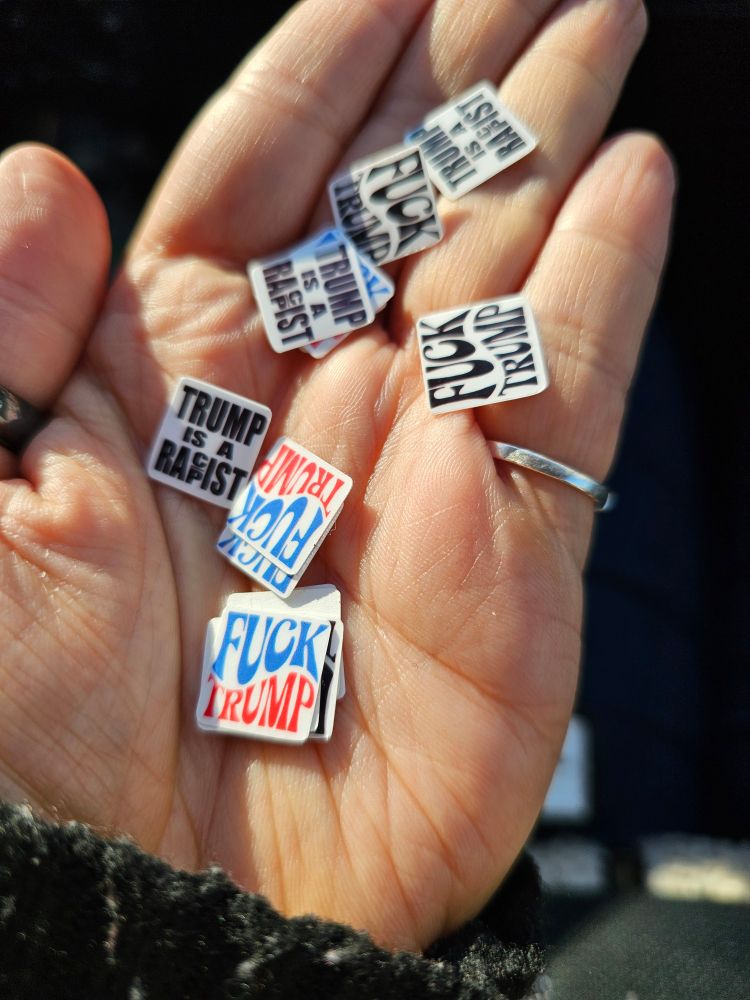 A wyt hand holding a bunch of tiny stickers that say Fuck Trump and Trump Is A Racist/Rapist