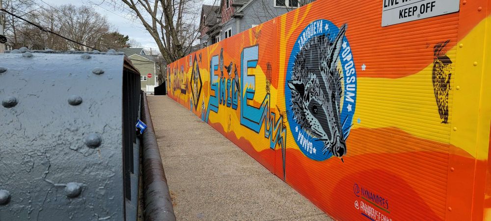 Pathway over rail line with new mural. Pretty orange, yellow, and red. A blue circle highlights the mighty opossum.