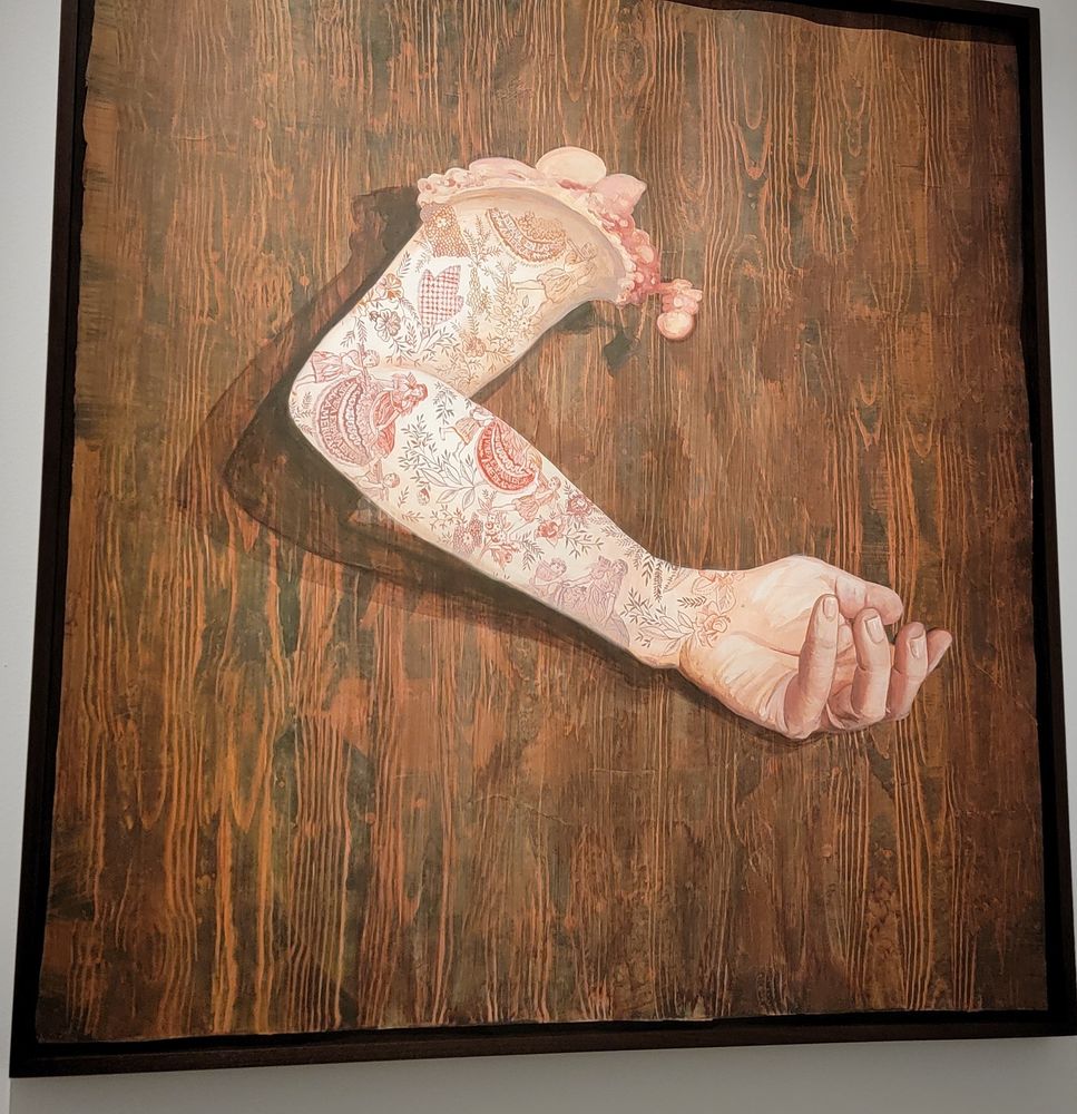 Firelei Báez - Retablo (españa en las Americas). A severed arm covered in delicately inked tattoos on a wooden background. The detail in her work is incredible