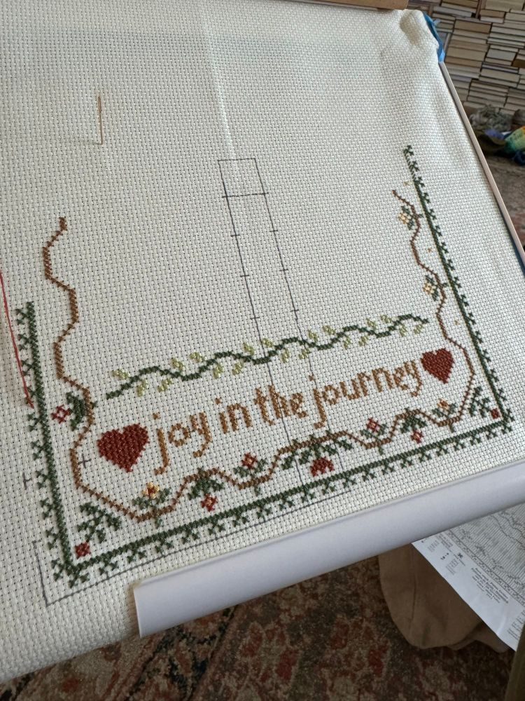 An in-progress cross stitch is mounted on a pvc holder. The words “joy in the journey” with red hearts on either end are visible at the bottom. There is a green vine above the words. There are red and yellow flowers around the border. The border is unfinished