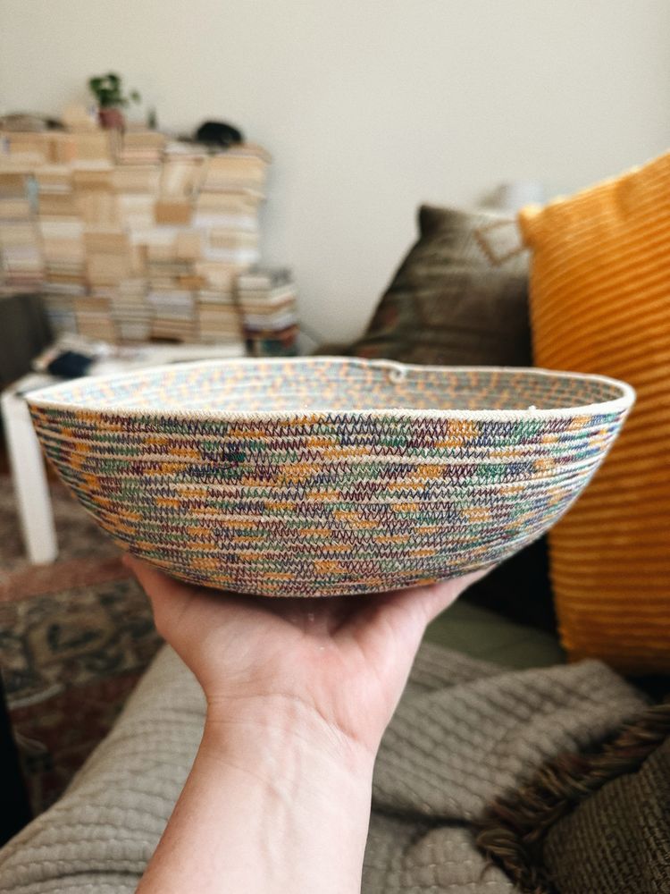 A white woman’s hand is holding a rope basket in the foreground, with a living room scene in the background. The basket is sewn with a zigzag stitch using teal, purple, and yellow multi-colored thread. 