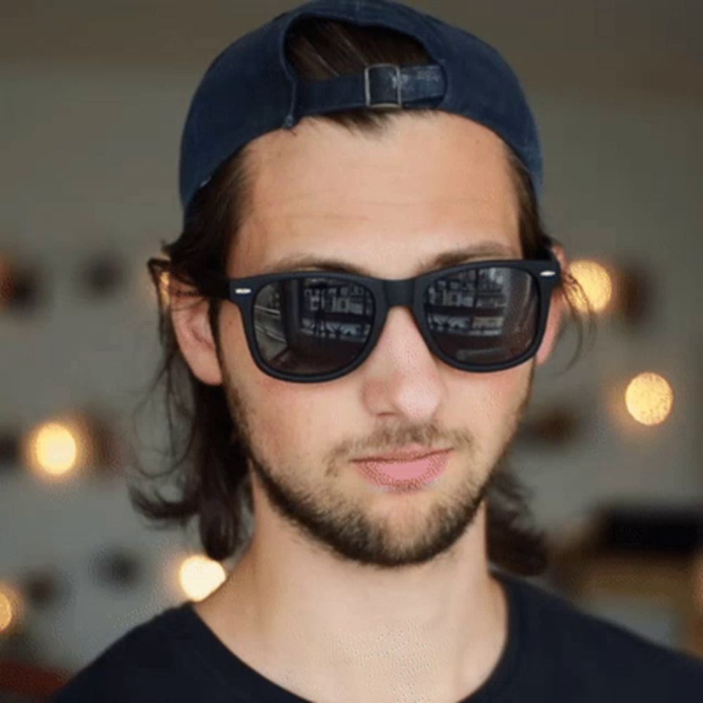 a man wearing sunglasses and a baseball cap has a reflection of a building in his sunglasses