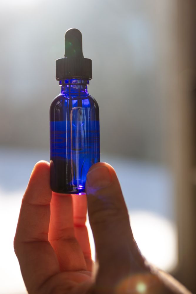 A dark blue bottle with a rubber dropper, held at the base by fingertips, backlit by warm light and a few small lens flares giving a dreamy, magical effect.