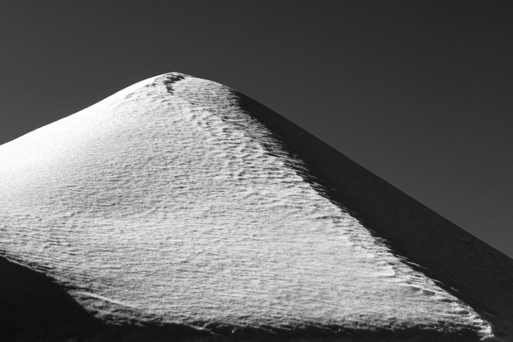 a (black and white) snowy rooftop appearing to be a very pyramidal mountain, with bright sunshine on the left face and dark shadow on the right