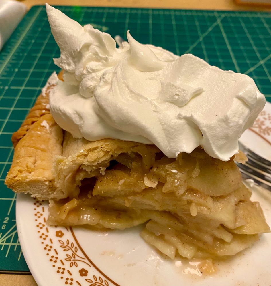 A slice of homemade apple pie overflowing with apple slices. A helping of whipped cream on top.