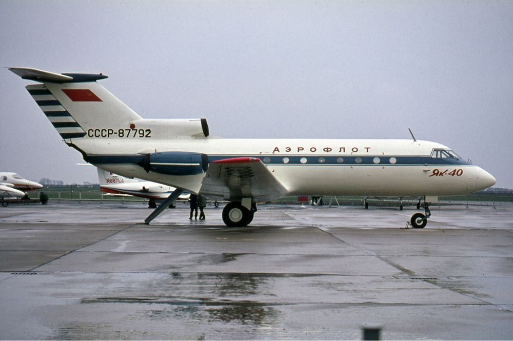 A Yakovlev Yak-40 airliner in Cold War era Aeroflot livery with it’s air stair down; viewed from the right side on a rainy day, several other Soviet airliners in the background. 