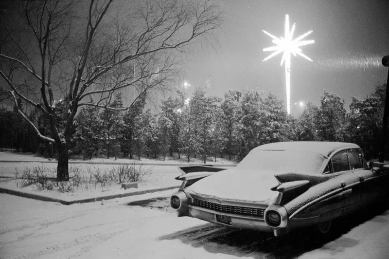 “Christmas, Kennedy Airport, 1967 by Joel Meyerowitz. A black and white image of a classic car covered in snow at a parking lot with a glowing Christmas star in the background. 