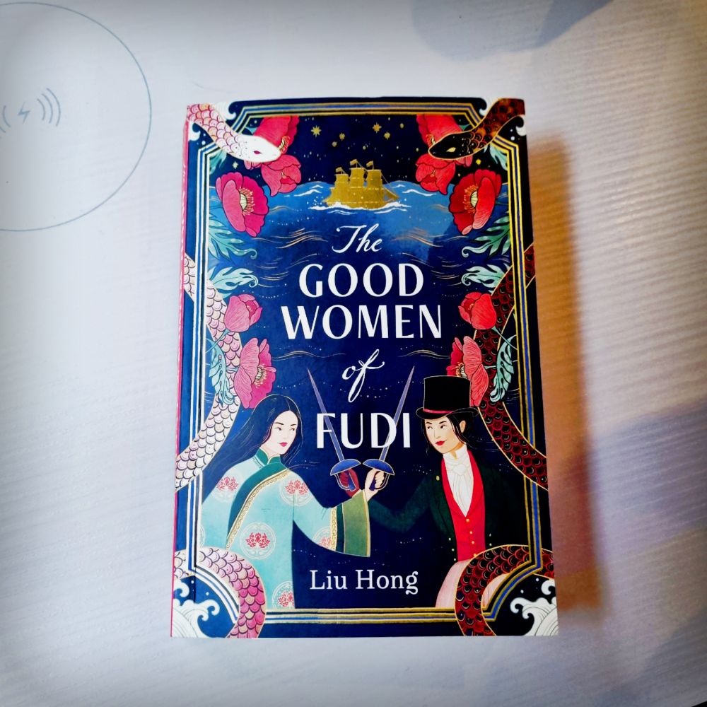 The book lying on a train table of fake cream wood. The cover has an illustration of two women, one in traditional Chinese dress, one in Elizabethan mens clothes with fencing swords crossed. The border has a black snake and a white snake, and red poppies inter twined. Off in the background is the silhouette of a tall ship foiled in gold. 