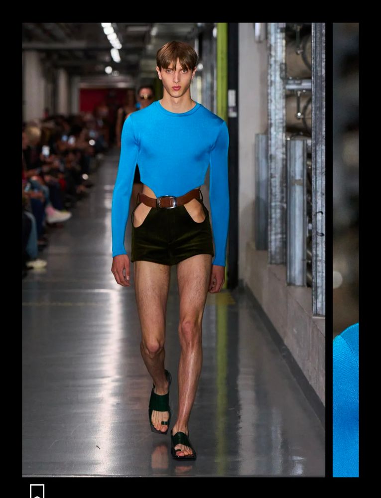 The opposite of the prior look, sort of, is the same space blue for a top, but it's skintight and covers the male model's whole torso and arms, and instead of khaki pants it's a little black skirt showing his hips, attached to the shiny space top. There's a nonfunctioning belt, and those goddamned clunky open toed shoes again 