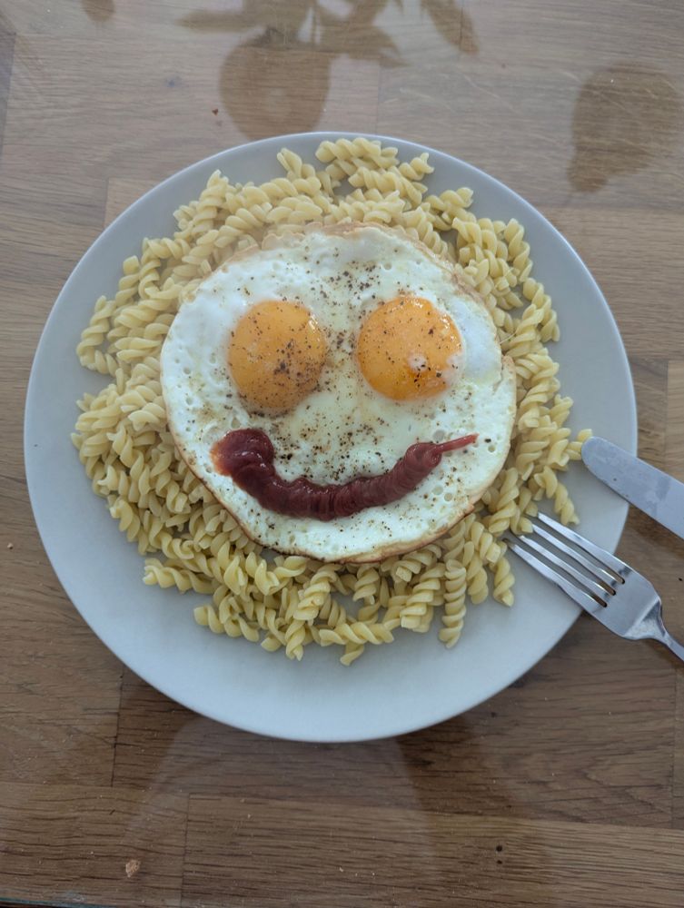 Plat de torsades dans une assiette blanche, surmontés de deux oeufs au plat de forme circulaire et d'un large sourire au ketchup, formant un smiley.